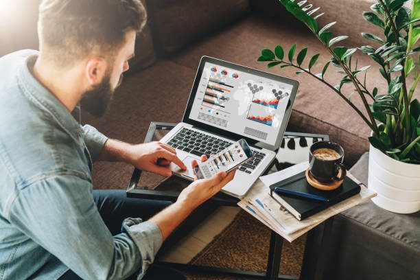 Young man, entrepreneur, and freelancer sits at home on the couch at the coffee table, uses a smartphone, working on a laptop with graphs, charts, and diagrams on screen. Online marketing, education, e-learning. Startup.