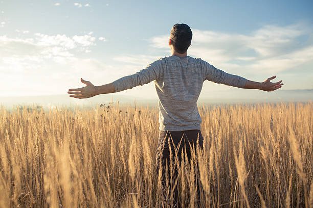 A young caucasian man standing in the middle of a prairie with his arms outstretched, enjoying a beautiful sunny day in nature.